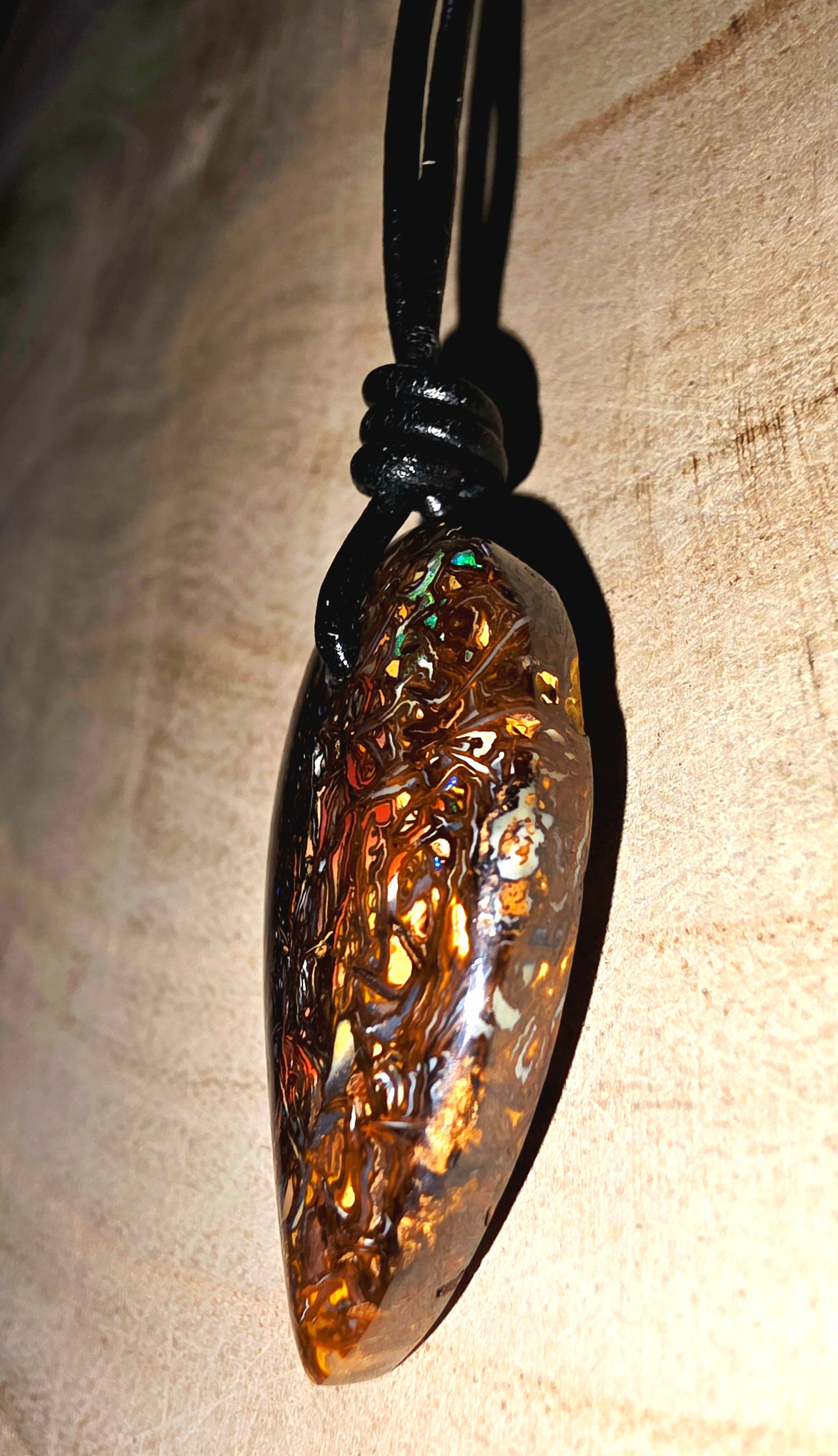 A teardrop-shaped opal pendant with intricate carved details, hanging from a black cord against a textured beige background.