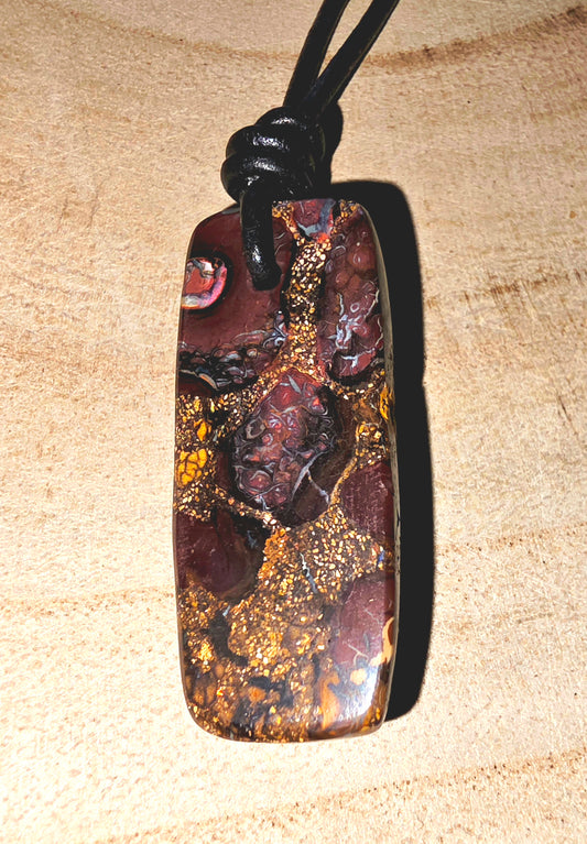 A polished Australian boulder stone pendant with a rich pattern of gold, brown, and dark red, suspended from a black cord on a light wood background.