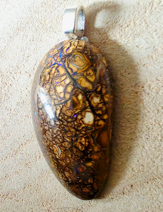 A teardrop-shaped pendant featuring a polished brown and gold mottled opal with dark web-like veining and small iridescent blue-purple flecks, suspended from a silver bail and photographed on a textured beige surface.
