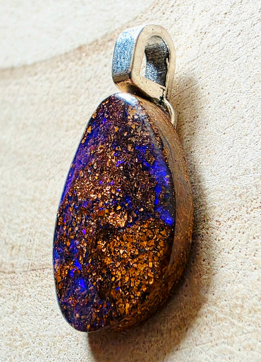 A teardrop-shaped pendant featuring a highly iridescent opal stone with shimmering gold and deep blue-purple flashes, set in a silver-toned metal bezel, resting on a textured beige surface.
