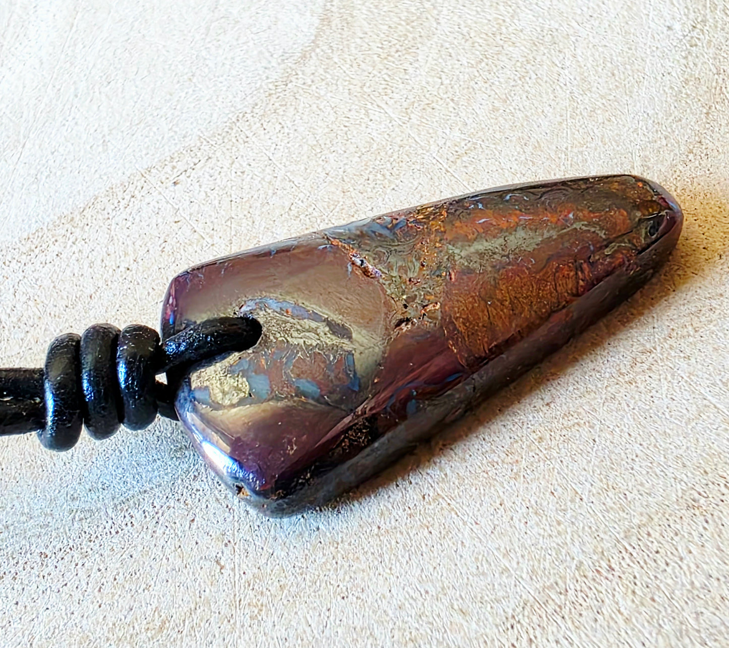 A polished natural opal pendant with a dark purple and metallic copper-brown marbled pattern, threaded on a black cord, resting on a textured beige fabric background.