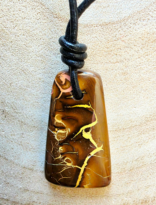 A polished brown stone pendant with gold veining, suspended from a black cord, set against a textured beige background.