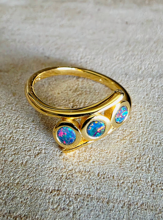 A gold ring featuring three round bezel-set iridescent opal stones, displayed on a textured beige background.