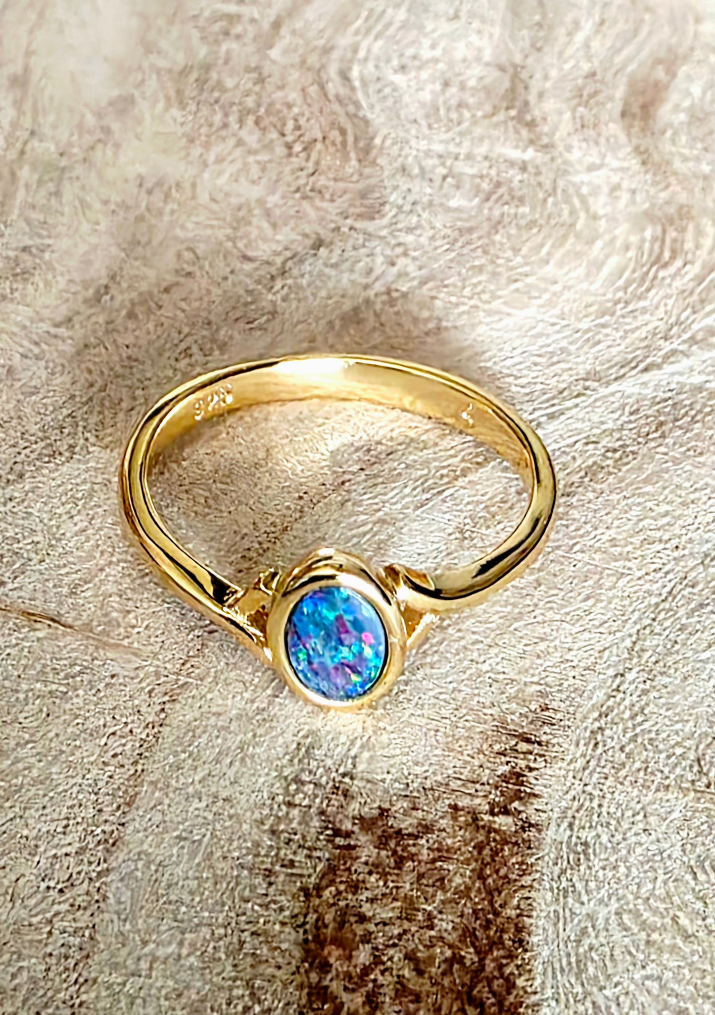 A gold ring with a round iridescent blue opal gemstone, resting on a textured beige background.