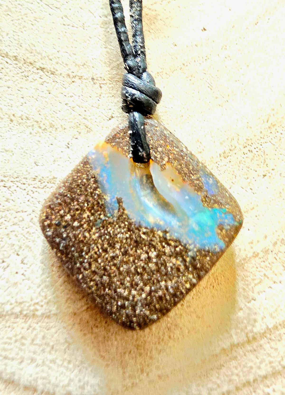 A square pendant necklace featuring a brown glittery textured base with a vibrant iridescent opal inlay of blue, white, and orange, suspended from a black cord on a textured beige background.