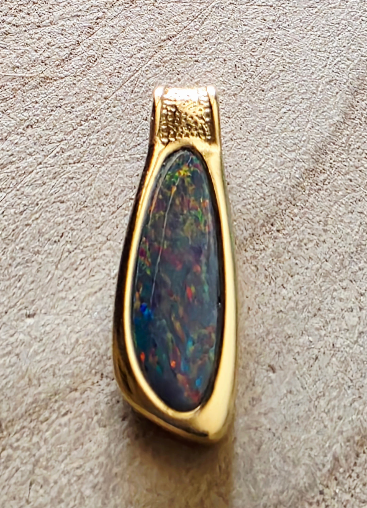 A gold pendant featuring a teardrop-shaped dark opal stone with vibrant multicoloured flashes, set against a textured beige background.