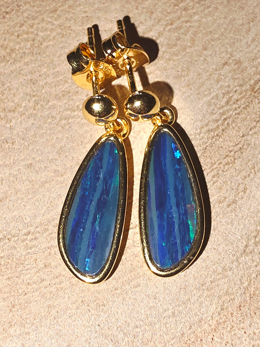A pair of gold drop earrings with teardrop-shaped blue opal gemstones, displayed on a textured beige surface.