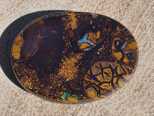 An oval-shaped polished opal gemstone with intricate patterns of dark brown, golden yellow, and iridescent flashes of blue, green, and orange, displayed on a textured beige stone surface.