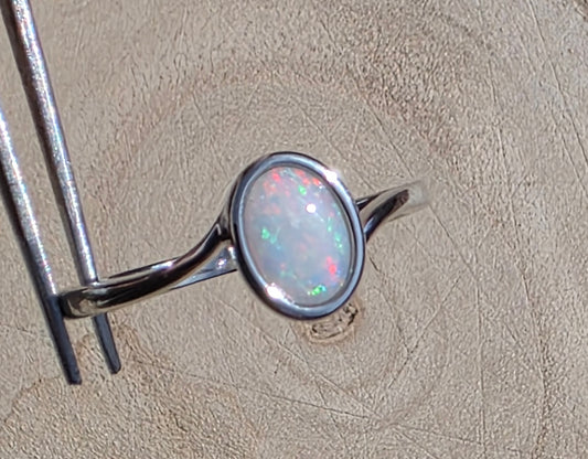 A sterling silver ring with an oval-cut white opal showing flashes of green and pink, held by metal tweezers against a textured beige background.