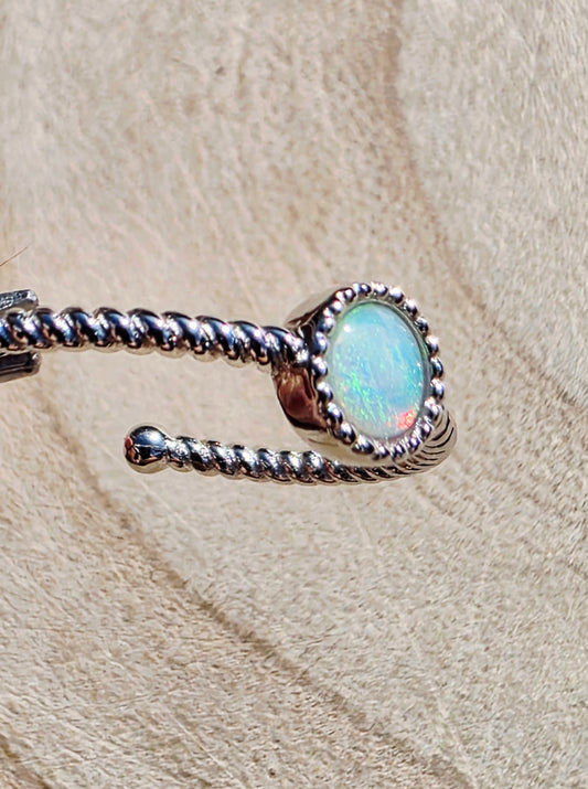 A silver-toned twisted rope ring with an oval iridescent opal stone, displayed on a textured beige surface.