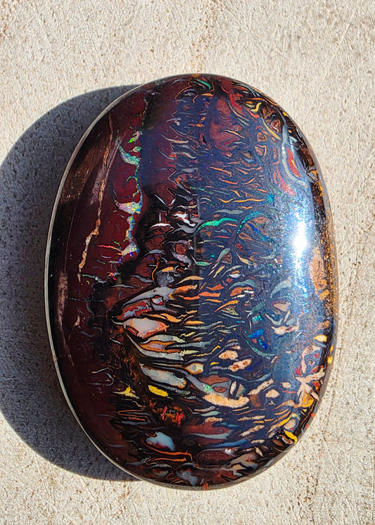 A polished oval opal gemstone with swirling iridescent patterns in red, brown, blue, green, and gold, photographed on a light beige textured surface.