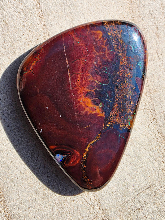 A polished natural opal stone with a teardrop shape, featuring a marbled pattern of deep red, orange, brown, and blue with gold flecks, set in a silver bezel on a light beige textured background.