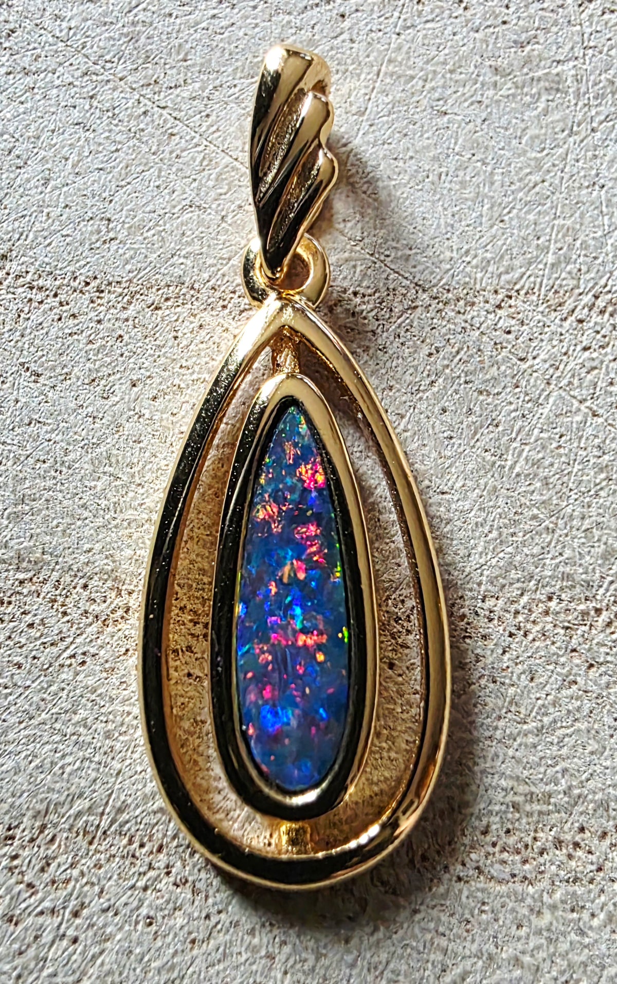 A gold teardrop pendant featuring a vibrant, multicoloured opal inlay, set against a textured light beige background.