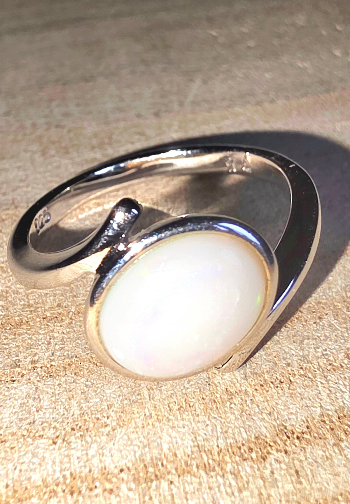 A sterling silver open band ring featuring a large, oval-shaped white opal gemstone, resting on a textured beige background.