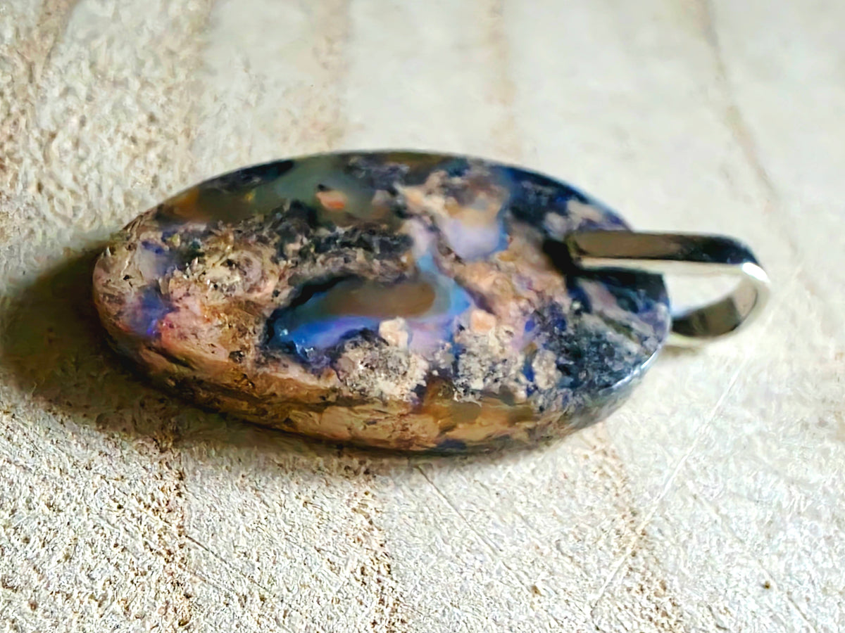 An oval pendant featuring a highly textured, marbled pattern of black, blue, brown, and beige, with a polished silver bail, resting on a textured beige background.
