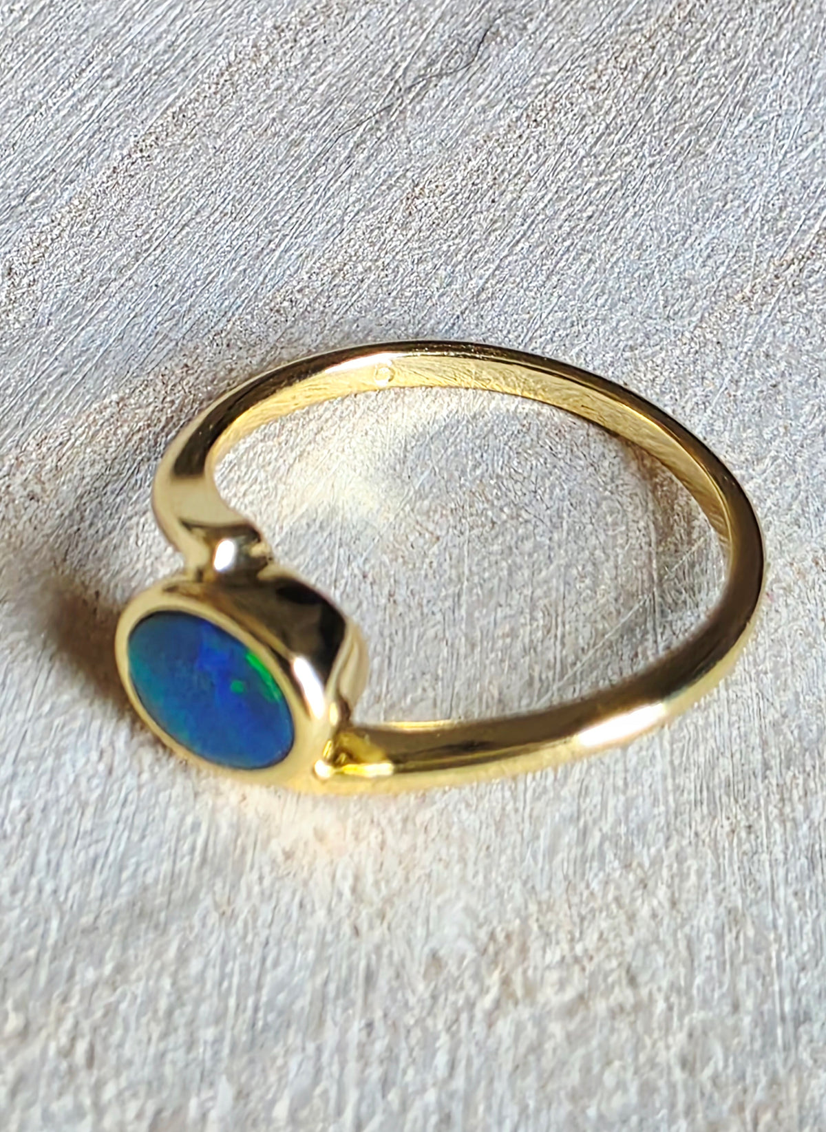 A gold ring featuring a round cabochon opal stone with vibrant blue and green iridescent colors, resting on a textured light beige surface.