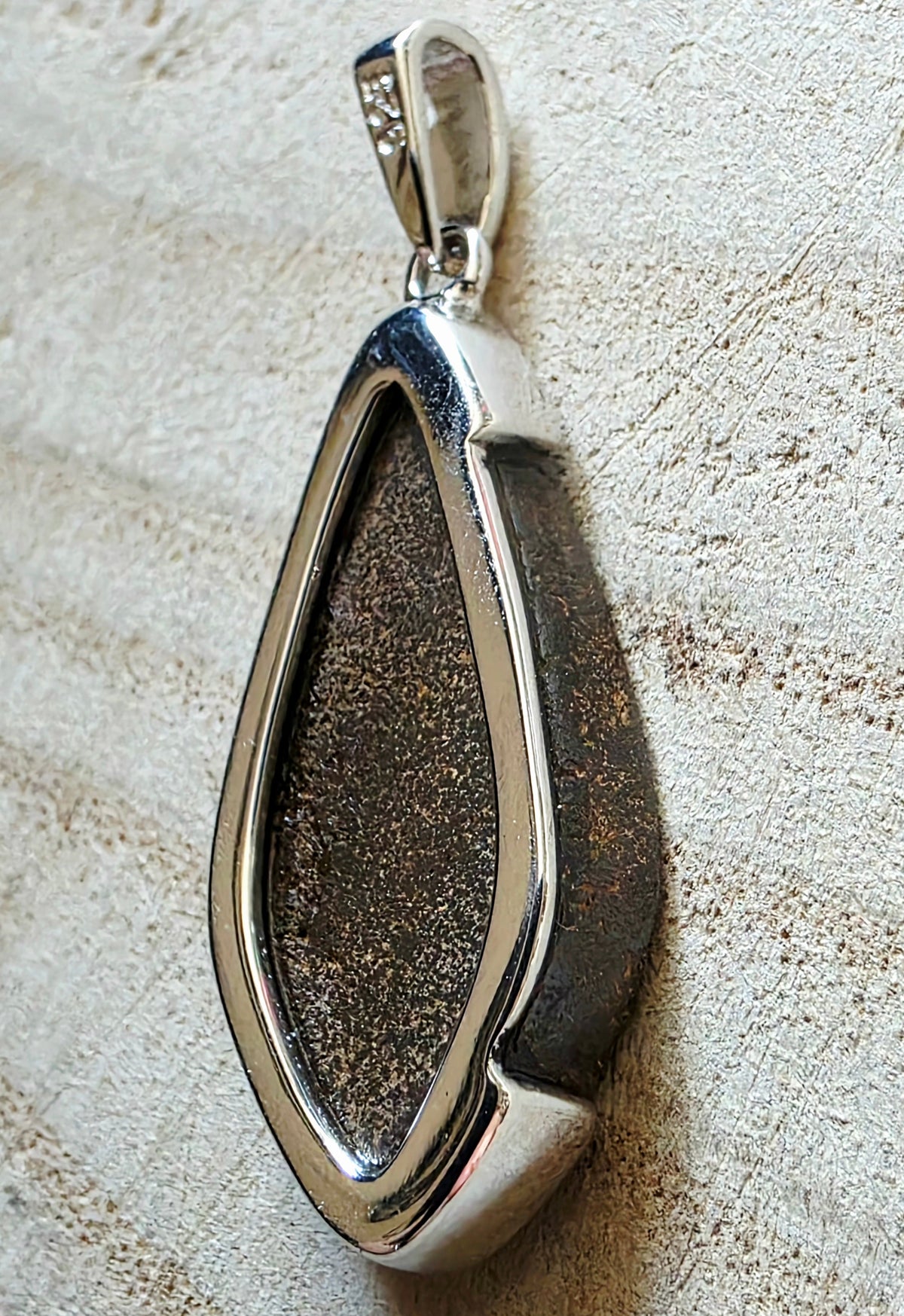 A teardrop-shaped pendant featuring a polished silver metal border and a textured, glittery dark brown centre opal, resting on a light beige textured surface.