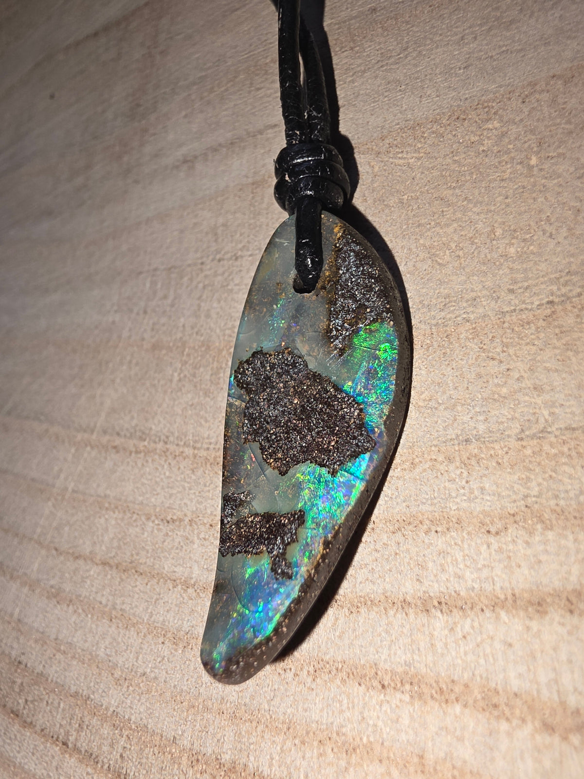 A teardrop-shaped Australian opal pendant with vibrant blue, green, and orange flashes, set in dark brown matrix and suspended from a black cord, resting on a light wood background.