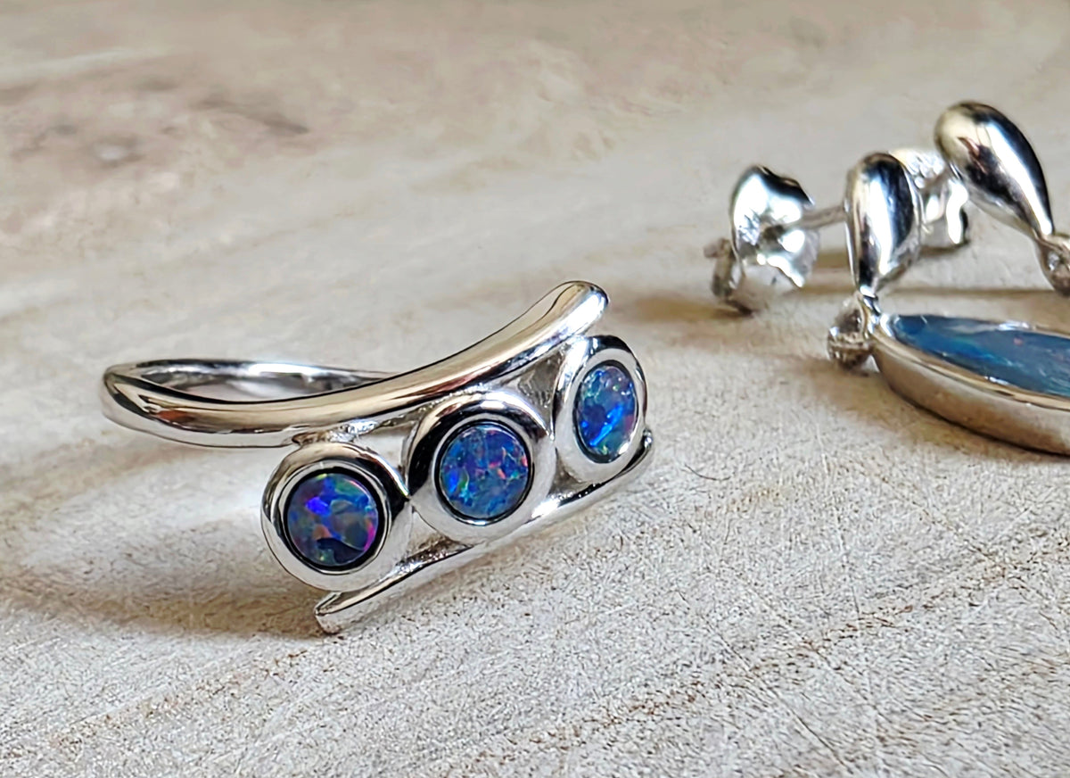 A sterling silver ring with three round blue opal gemstones, displayed on a textured beige surface next to matching silver earrings.