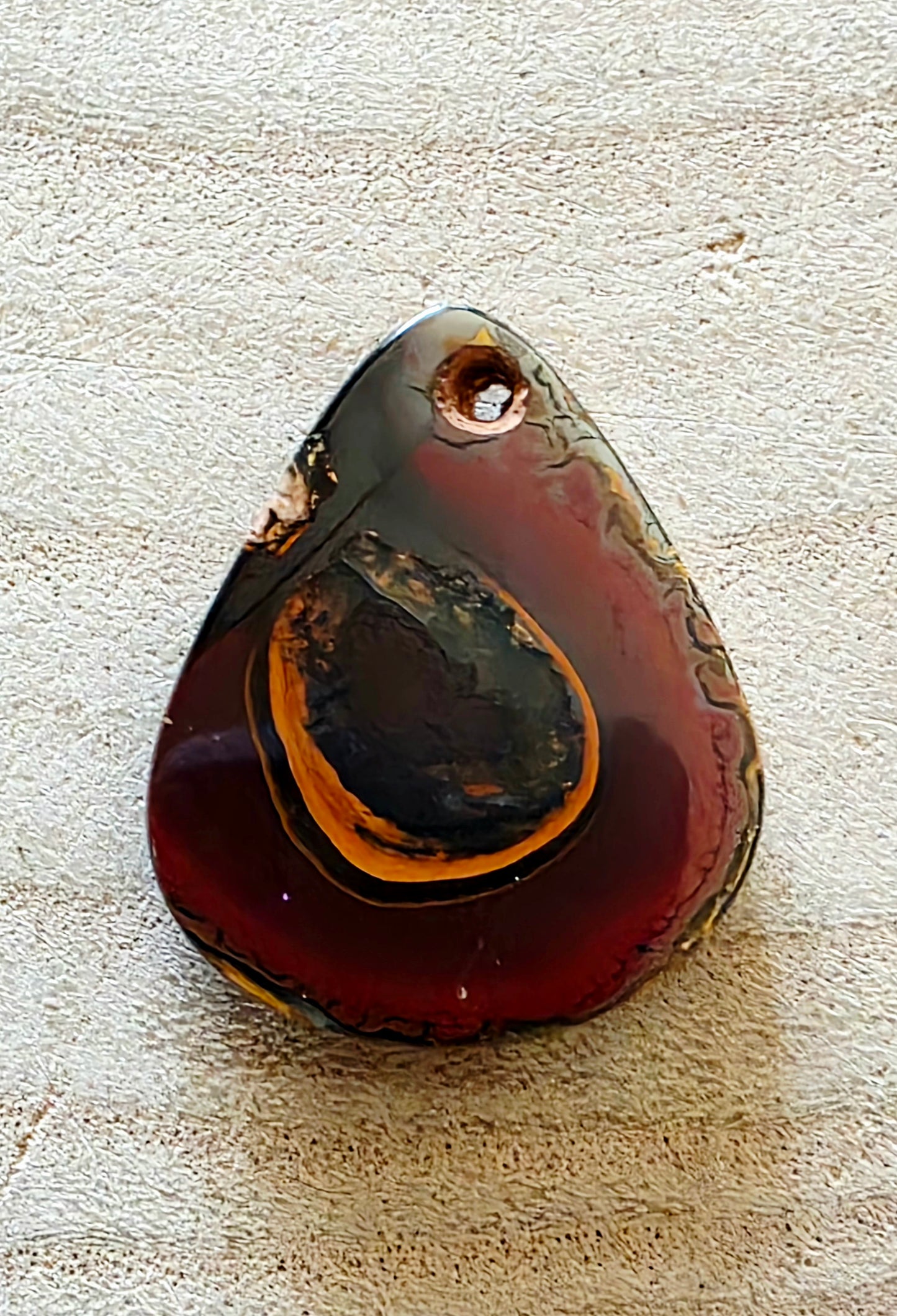 Teardrop-shaped pendant with a dark brown and black colour on a textured beige surface