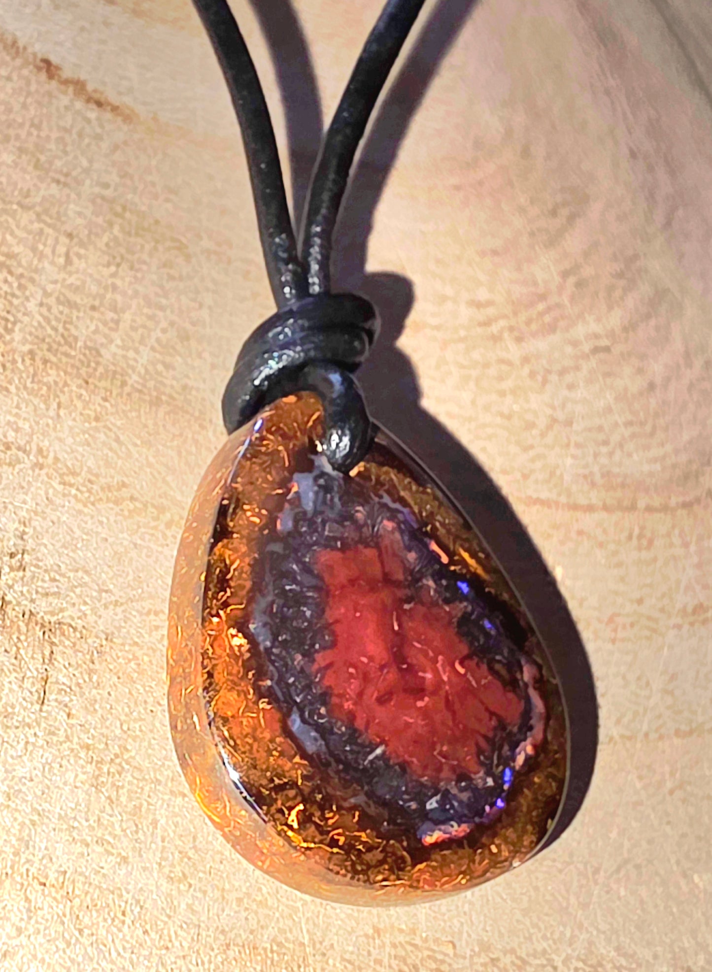 Teardrop-shaped pendant with a red and brown colour pattern on a black cord, against a wooden background.