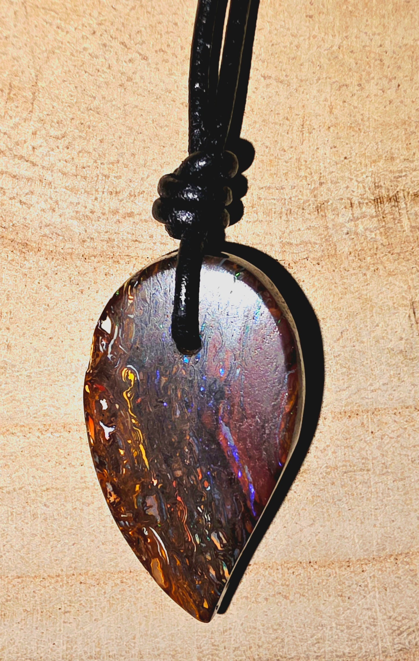 A teardrop-shaped iridescent opal pendant on a black cord, displayed against a light wood background.