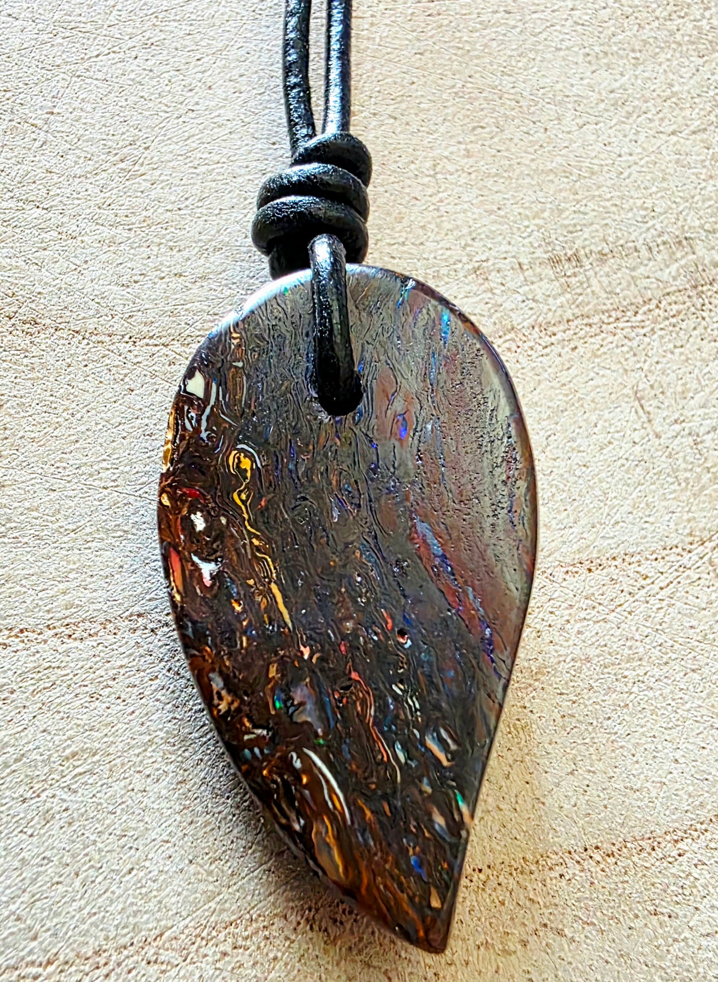 A teardrop-shaped pendant made of iridescent dark brown wood with shimmering gold, blue, and orange inclusions, hanging from a black cord on a light beige textured background.