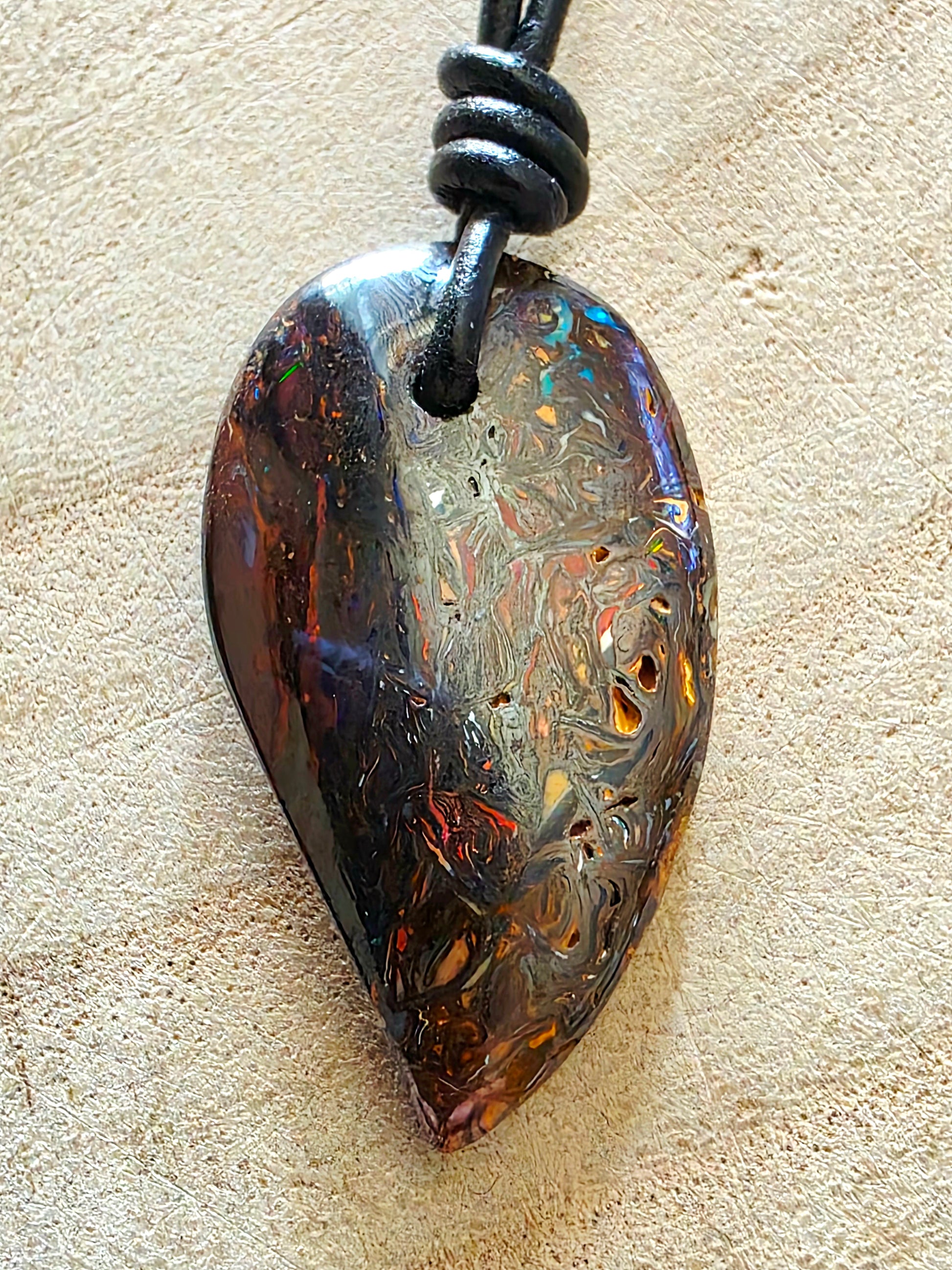 A teardrop-shaped pendant made of iridescent opal with swirling brown, gold, and blue patterns, suspended from a black cord on a textured beige surface.