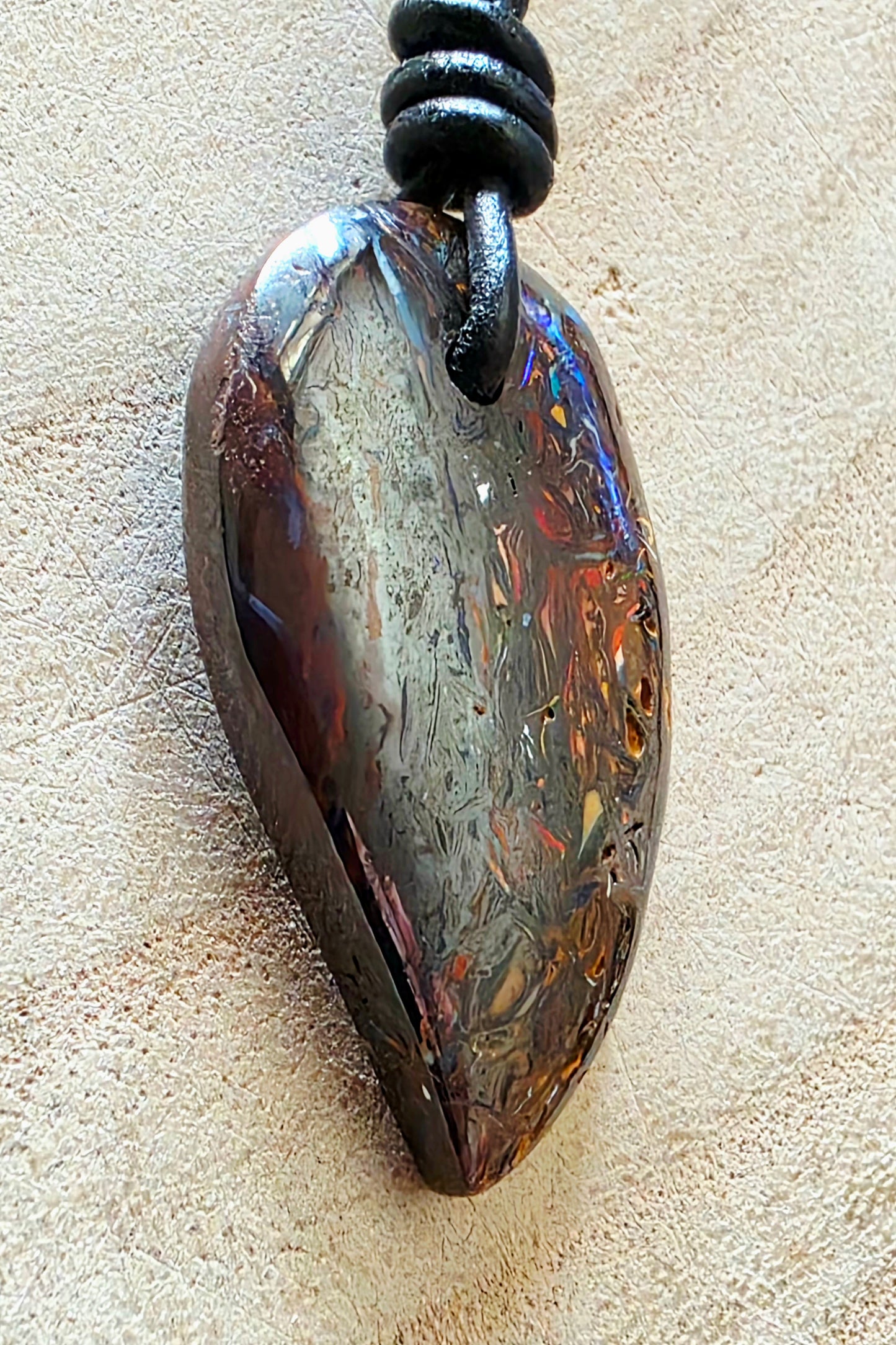 A teardrop-shaped iridescent stone pendant with swirling brown, gold, and blue hues, suspended from a black cord on a textured beige surface.