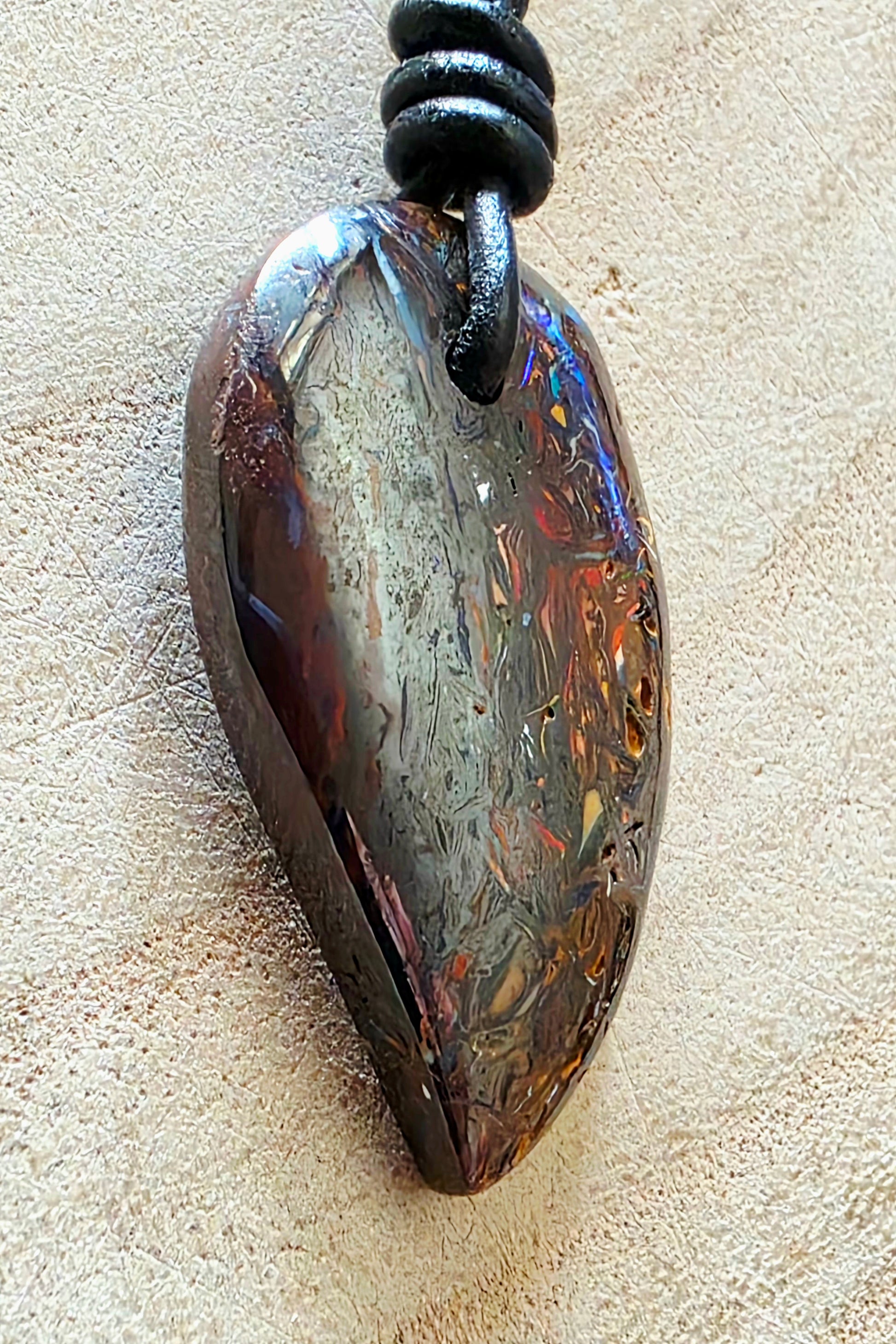 A teardrop-shaped iridescent stone pendant with swirling brown, gold, and blue hues, suspended from a black cord on a textured beige surface.