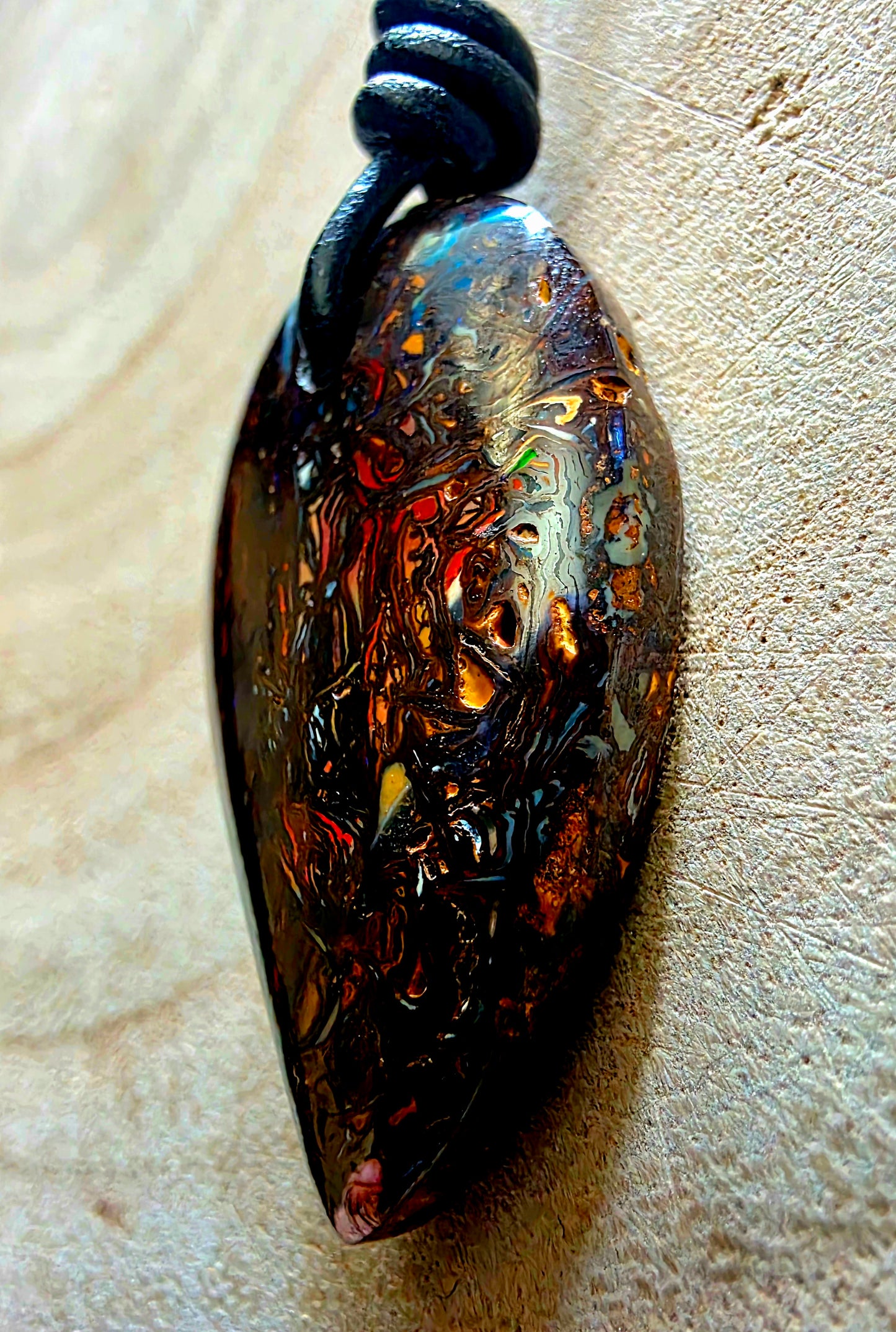 A teardrop-shaped pendant with a glossy, marbled pattern of black, copper, brown, and iridescent blue-gold tones, suspended from a black cord on a textured beige surface.
