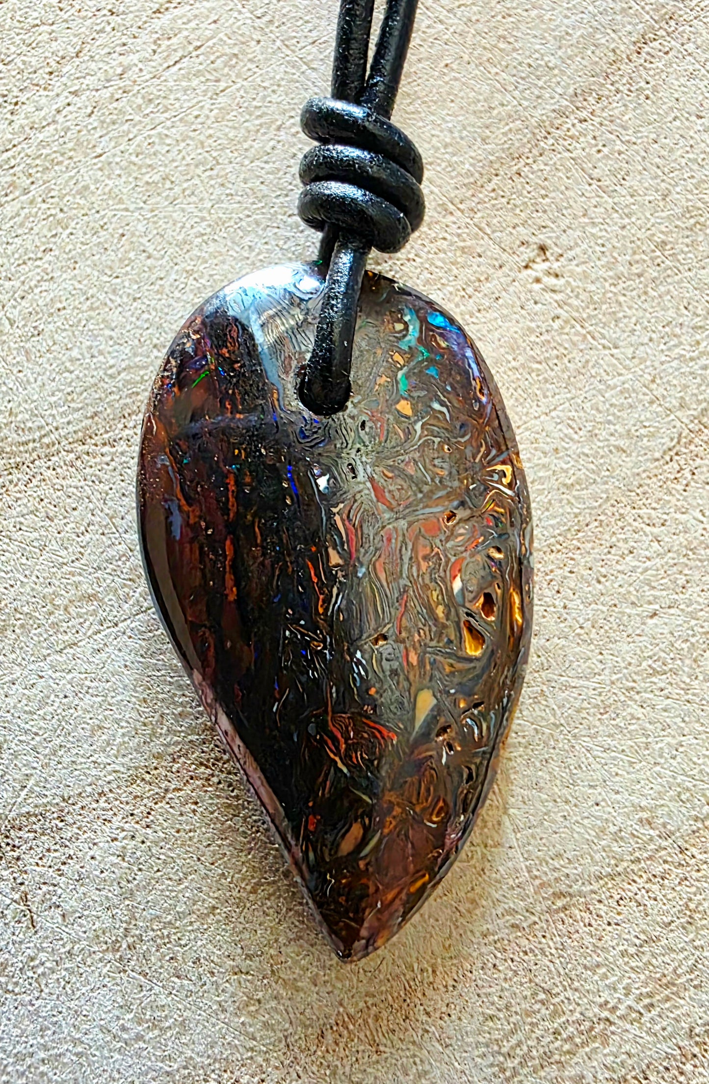 A teardrop-shaped iridescent opal pendant necklace on a black cord, displayed on a textured beige surface.