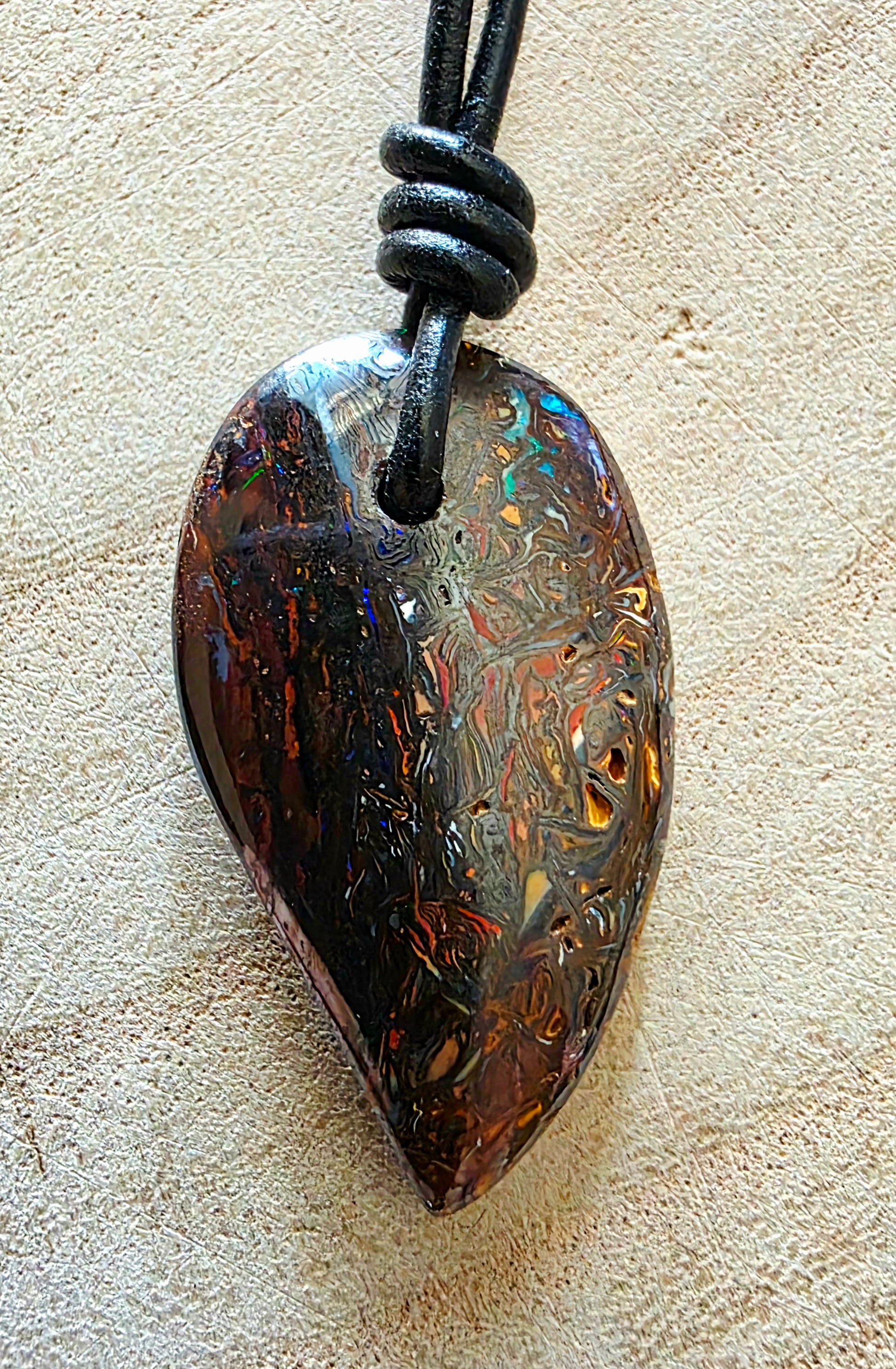 A teardrop-shaped iridescent opal pendant necklace on a black cord, displayed on a textured beige surface.