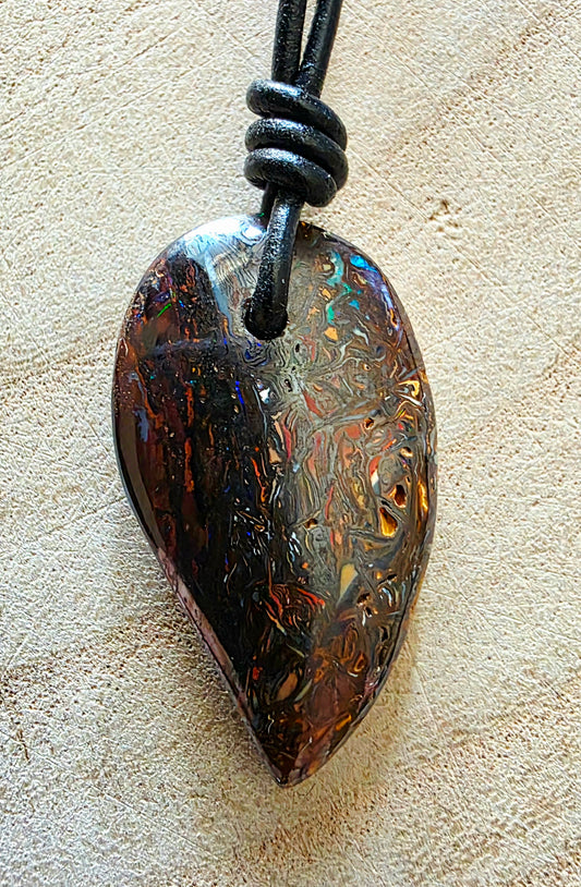 A teardrop-shaped iridescent opal pendant necklace on a black cord, displayed on a textured beige surface.