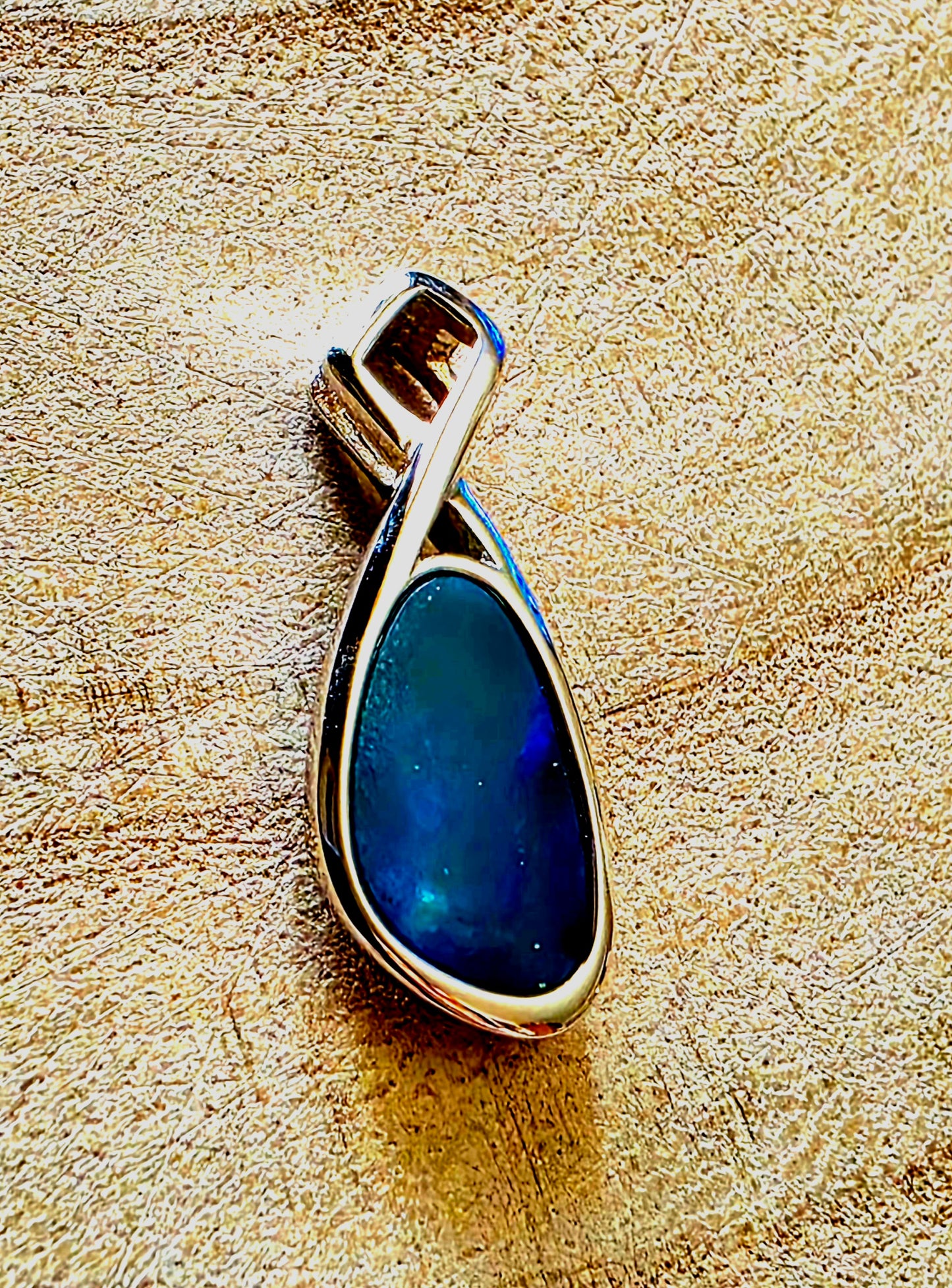A teardrop-shaped pendant featuring a deep blue iridescent stone in a polished silver bezel setting, displayed on a textured beige surface.