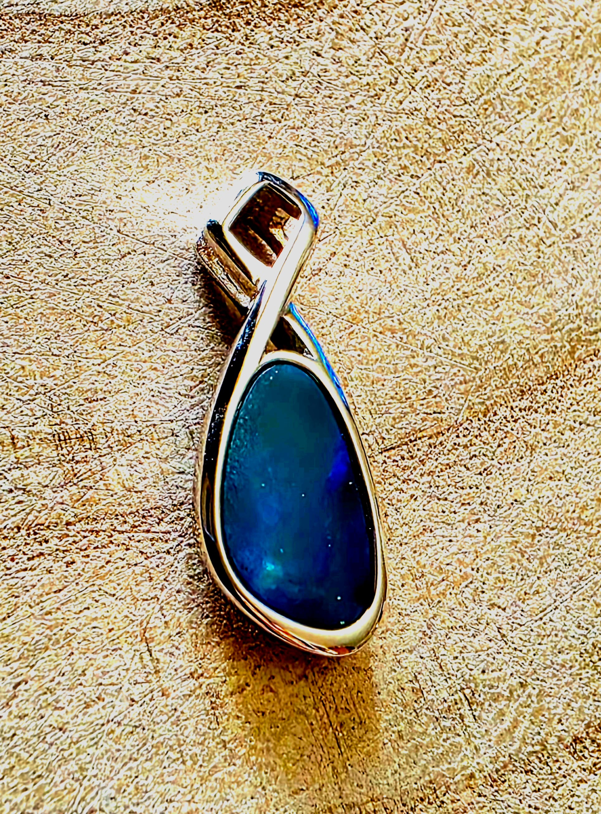 A teardrop-shaped pendant featuring a deep blue iridescent stone in a polished silver bezel setting, displayed on a textured beige surface.