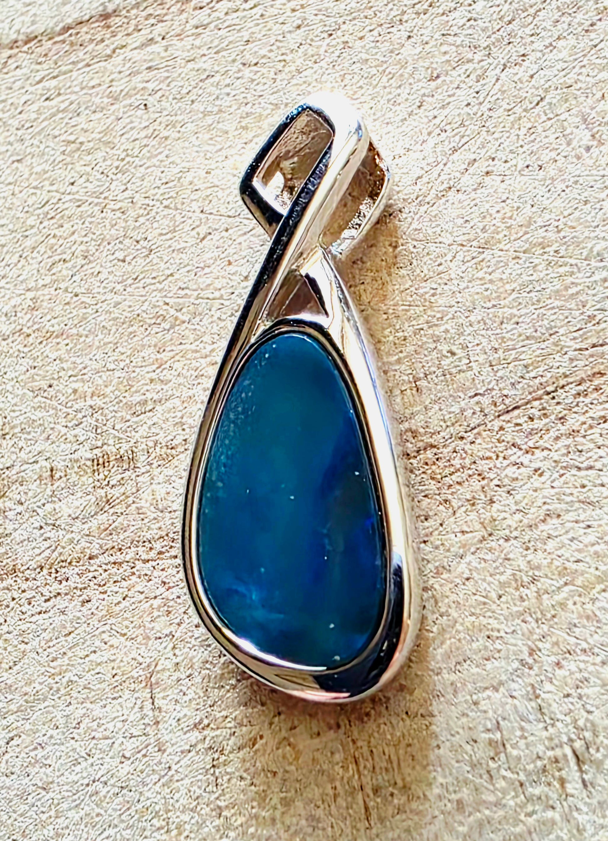 A teardrop-shaped pendant featuring a smooth, polished dark blue stone in a silver-toned metal setting, displayed on a textured beige surface.