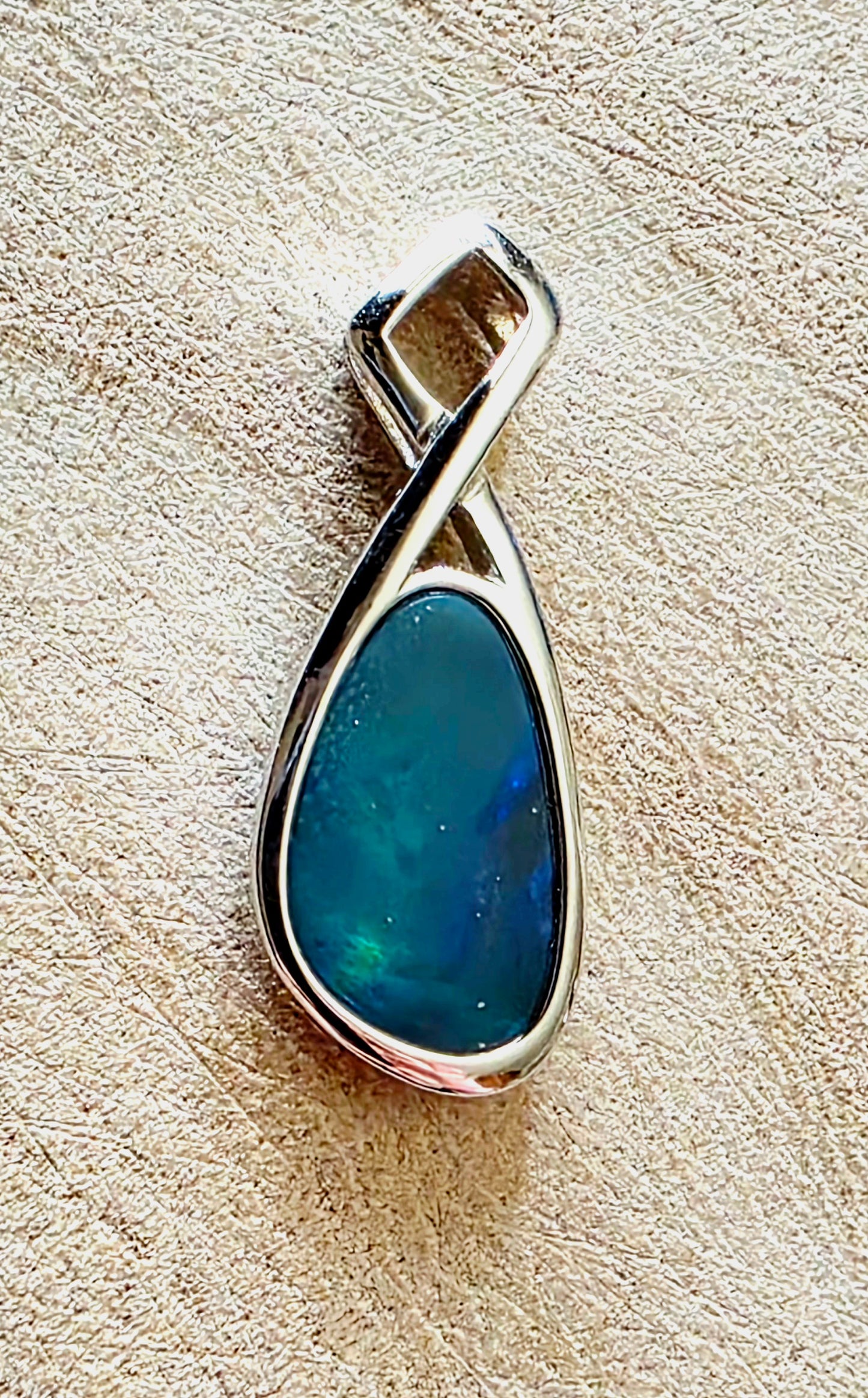 A silver teardrop pendant featuring a blue-green iridescent opal stone, displayed on a textured beige surface.
