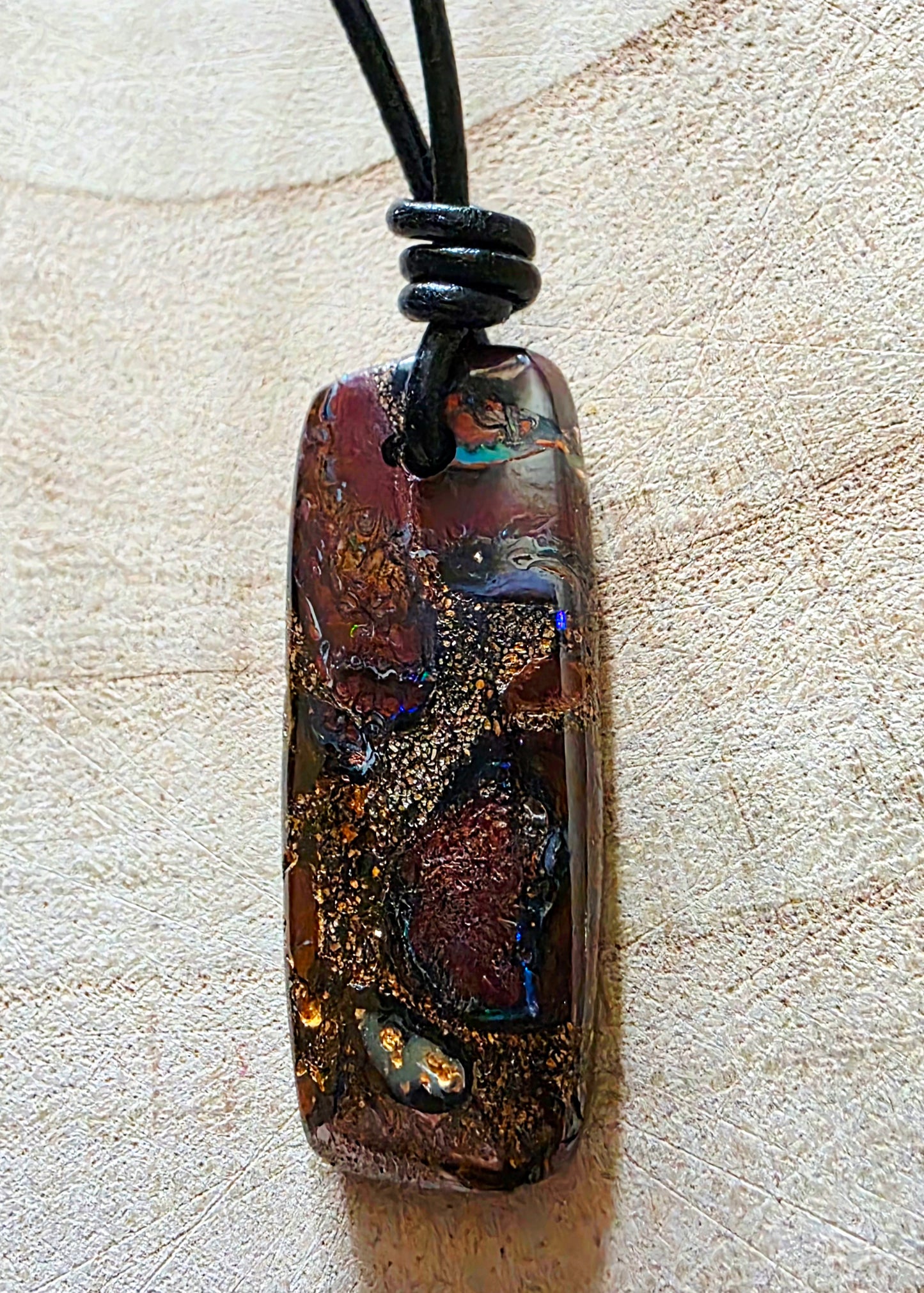 A rectangular pendant necklace with a black cord, featuring a marbled pattern of dark brown, black, and shimmering gold flecks, resting on a textured beige surface.