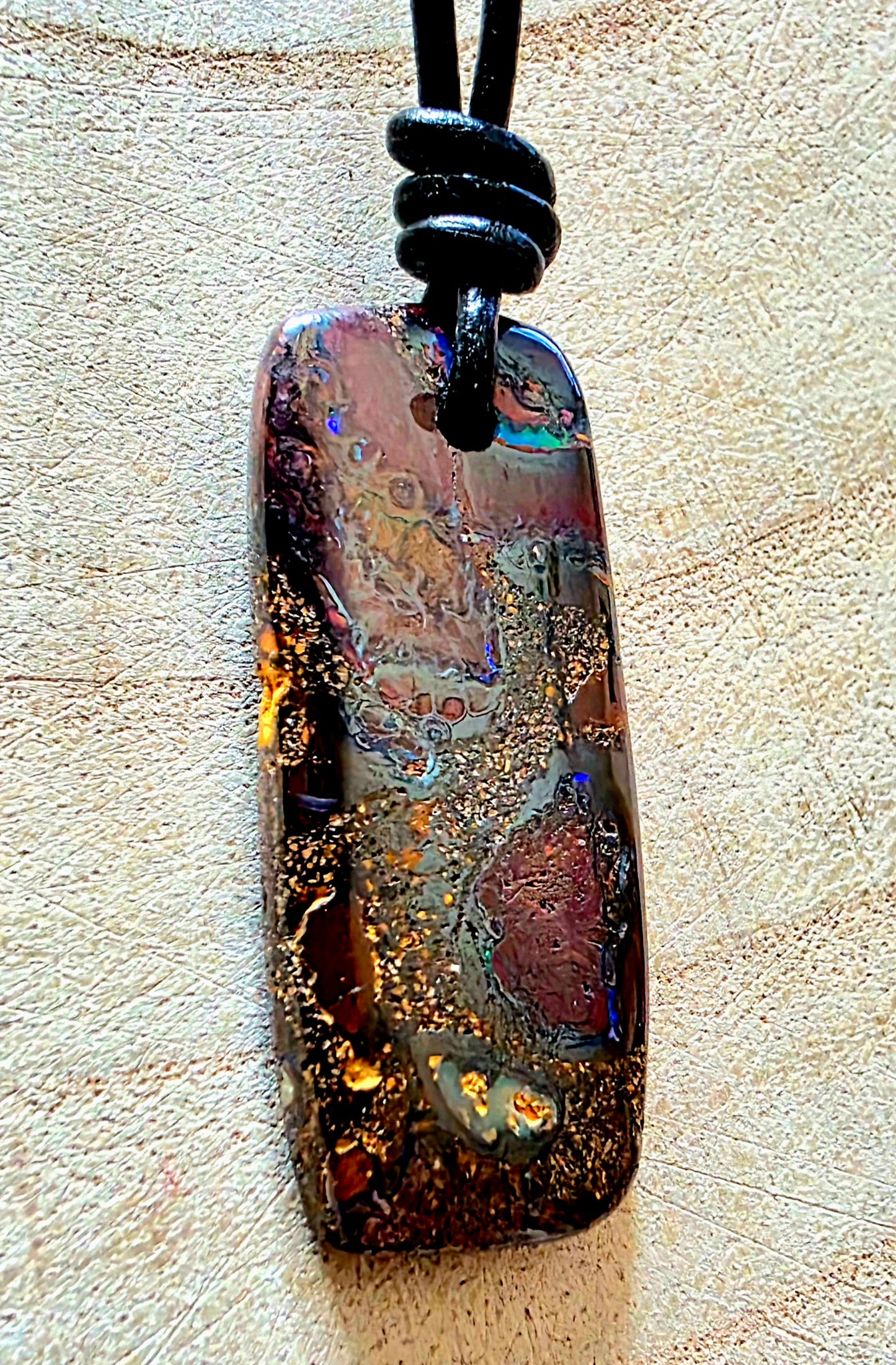 A rectangular polished opal pendant necklace with a black cord, featuring a highly iridescent play-of-colour pattern with gold flecks, resting on a textured beige surface.
