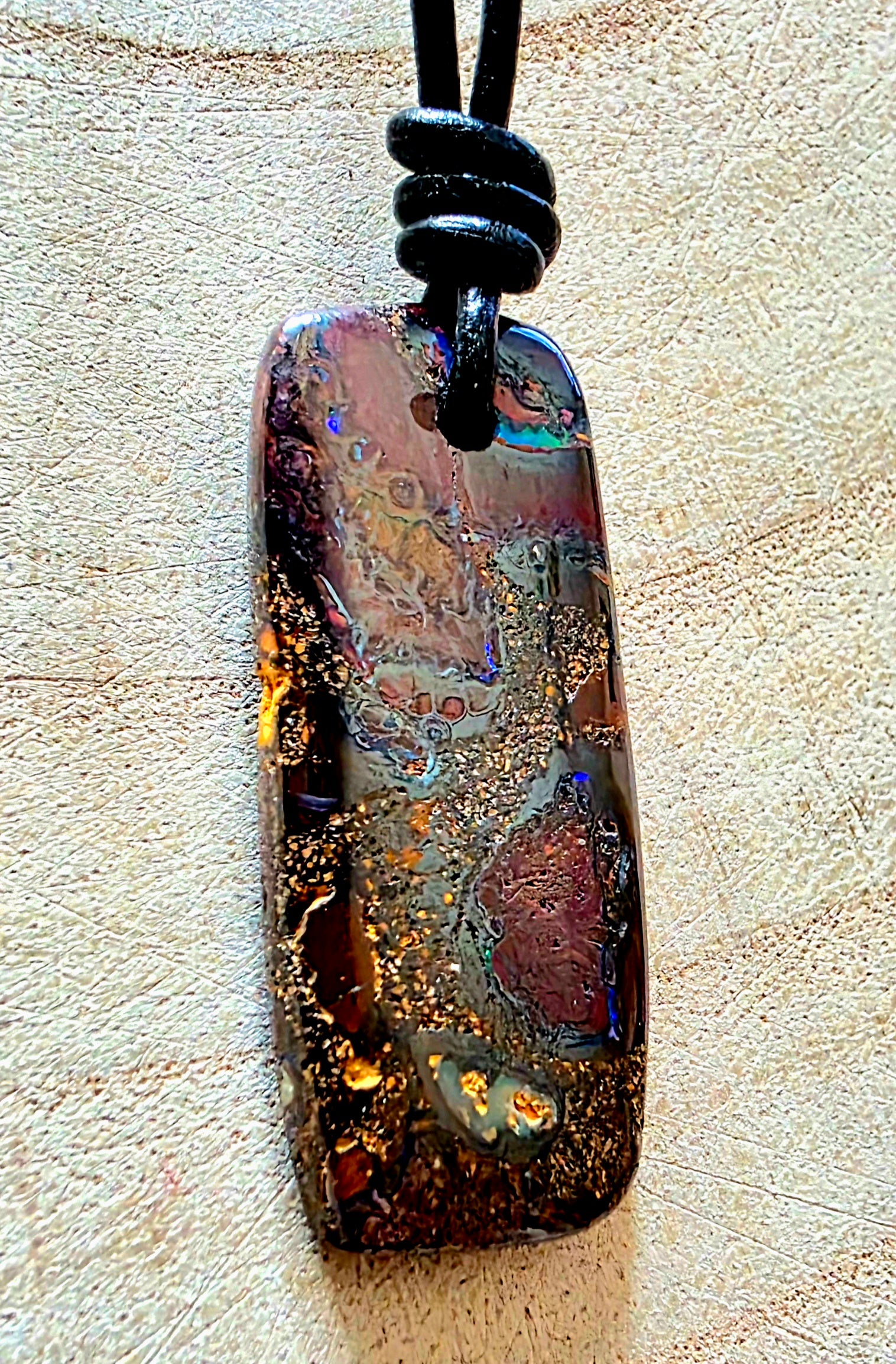 A rectangular polished opal pendant necklace with a black cord, featuring a highly iridescent play-of-colour pattern with gold flecks, resting on a textured beige surface.