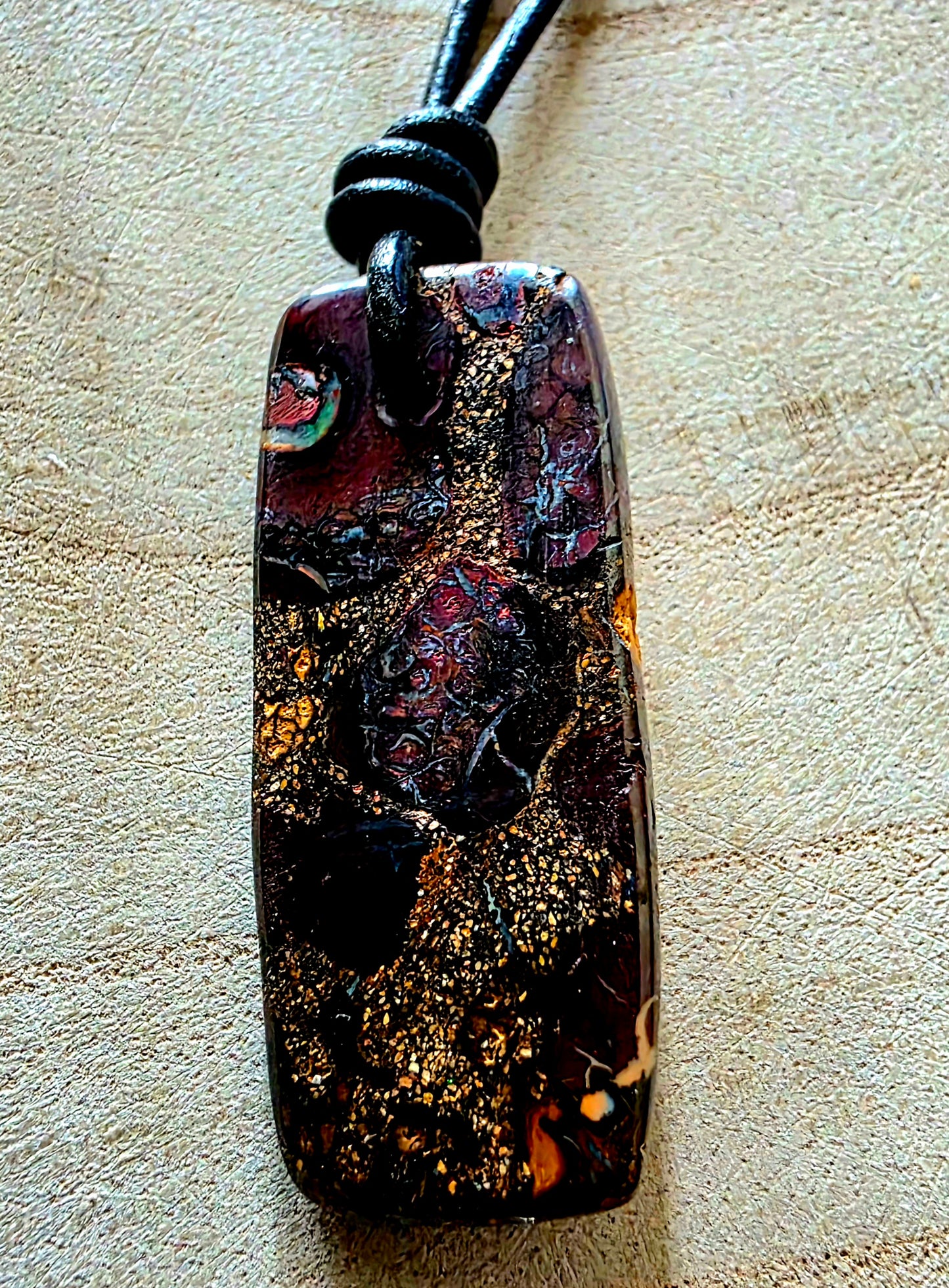 A rectangular pendant necklace with a glossy, marbled finish in black, gold, and deep red-brown tones, suspended from a black cord on a textured beige background.