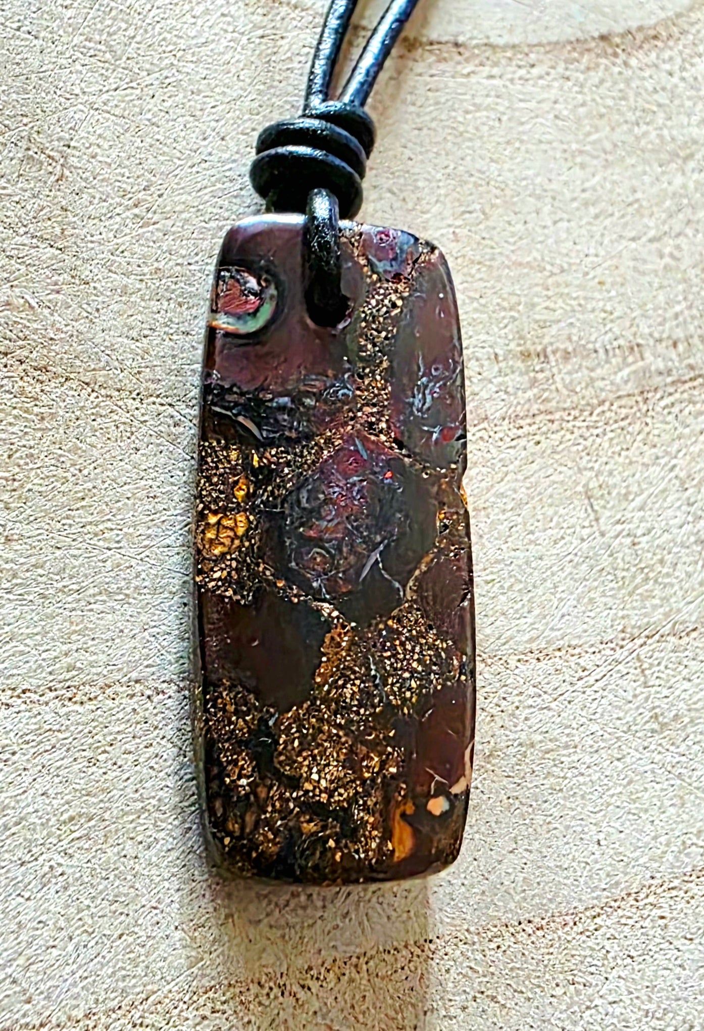 A dark brown and black opal pendant with gold flecks, suspended on a black cord, displayed on a light wood background.