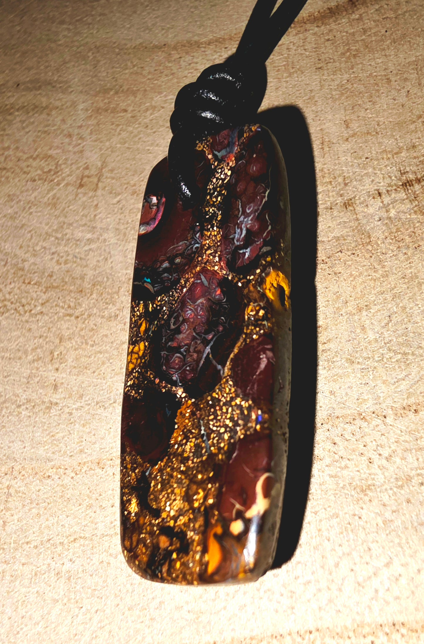 A polished dark brown and black opal stone pendant with prominent gold flecks, suspended from a black cord, displayed on a textured beige surface.