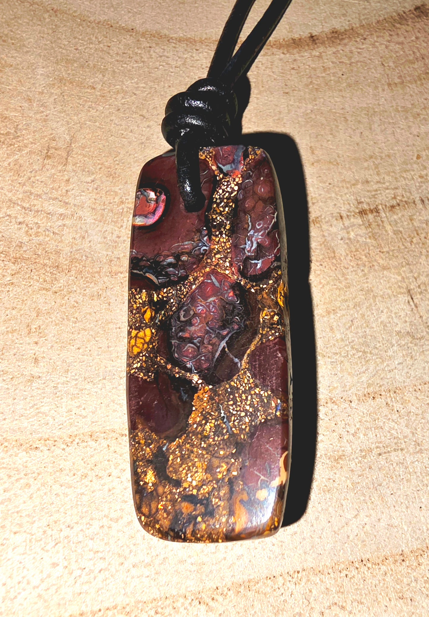 A polished Australian boulder stone pendant with a rich pattern of gold, brown, and dark red, suspended from a black cord on a light wood background.