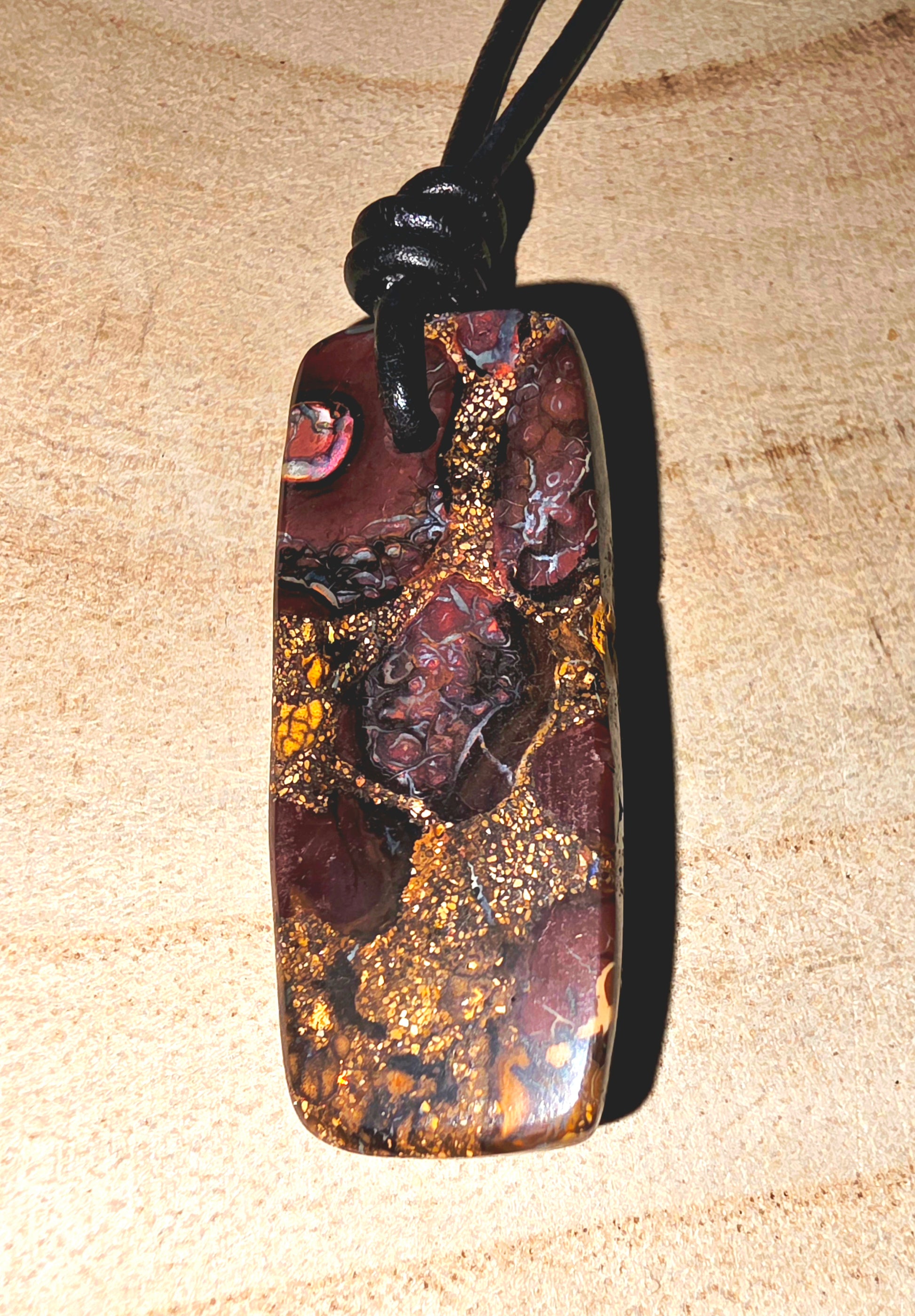 A polished Australian boulder stone pendant with a rich pattern of gold, brown, and dark red, suspended from a black cord on a light wood background.