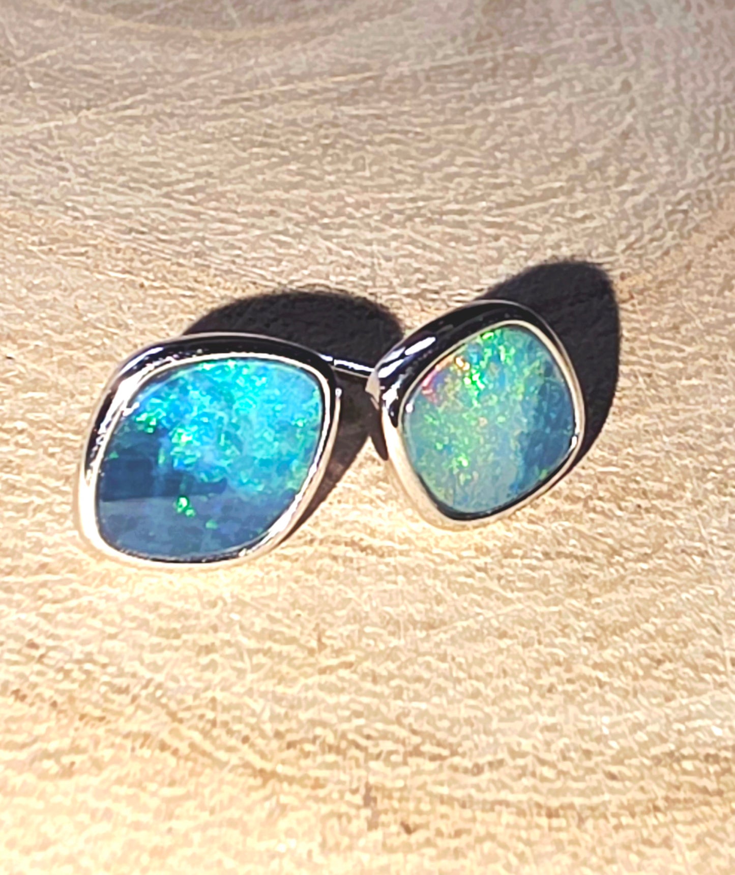 A pair of silver stud earrings with rounded square-cut blue and green iridescent opal stones, resting on a textured beige surface.