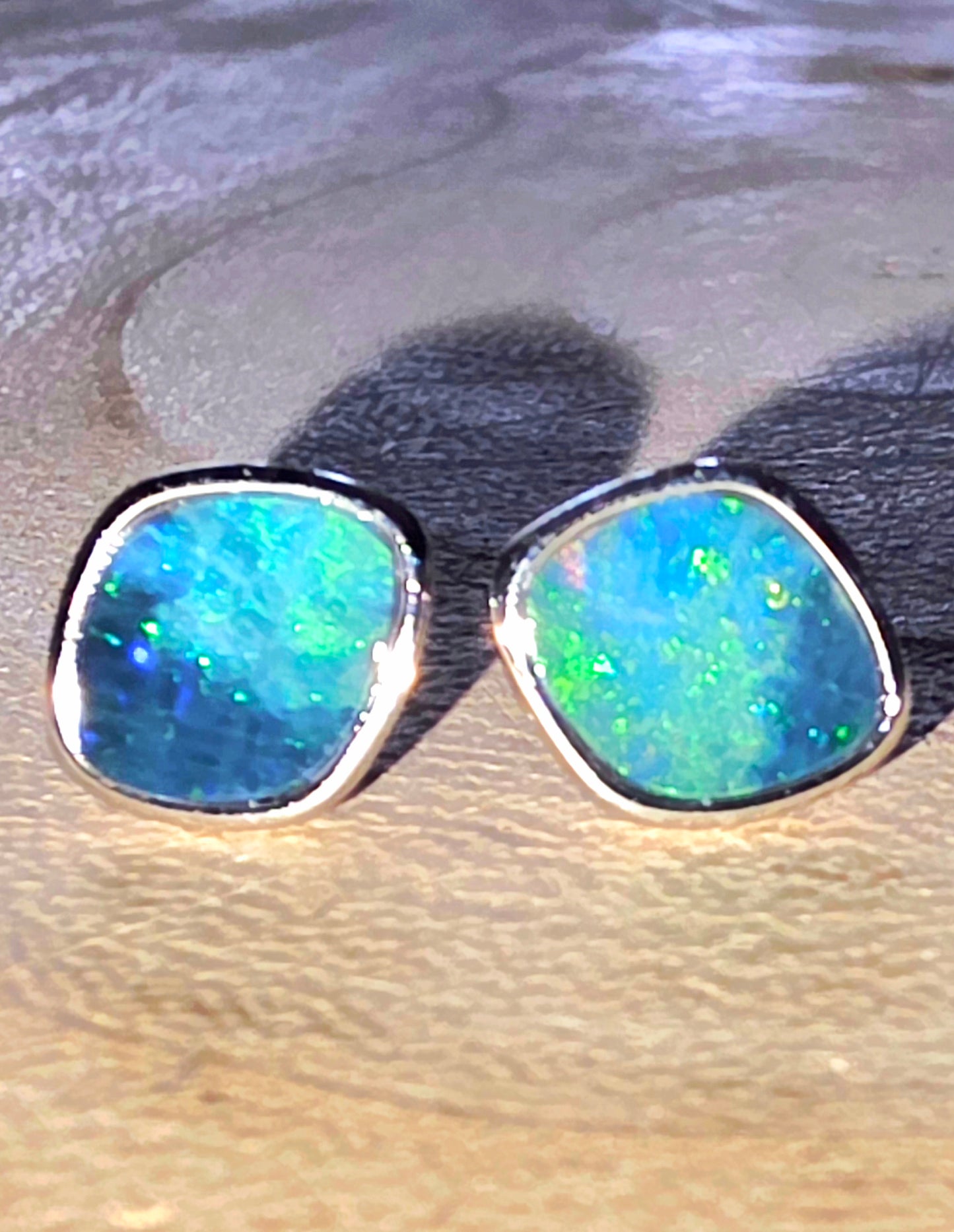 A pair of silver stud earrings with irregular square-shaped blue and green opal gemstones, resting on a textured beige surface.