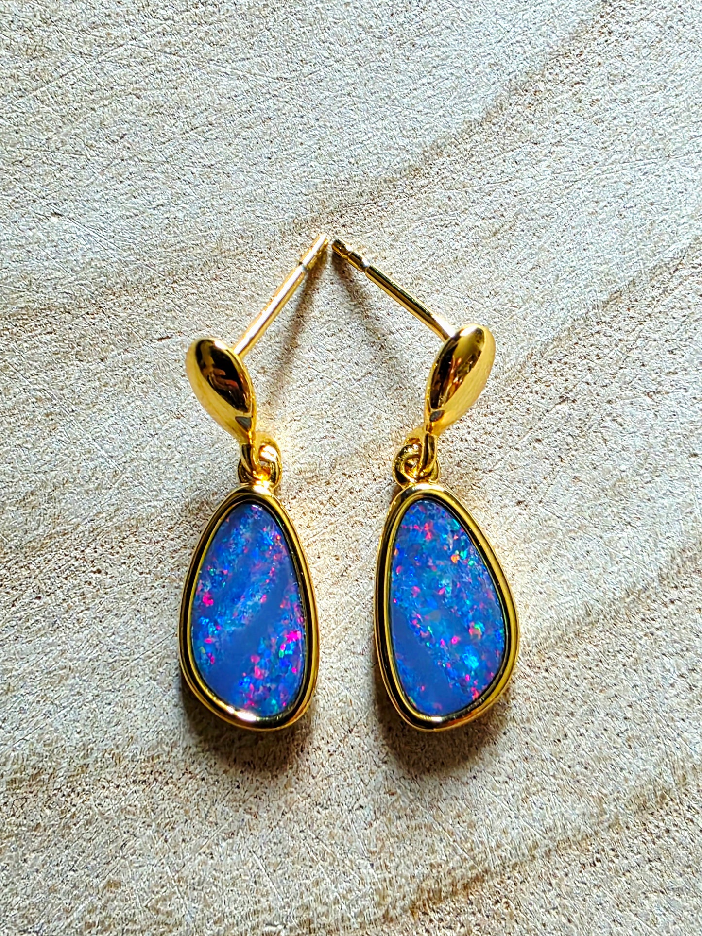 A pair of gold drop earrings with teardrop-shaped blue opal gemstones, displayed on a textured beige fabric background.