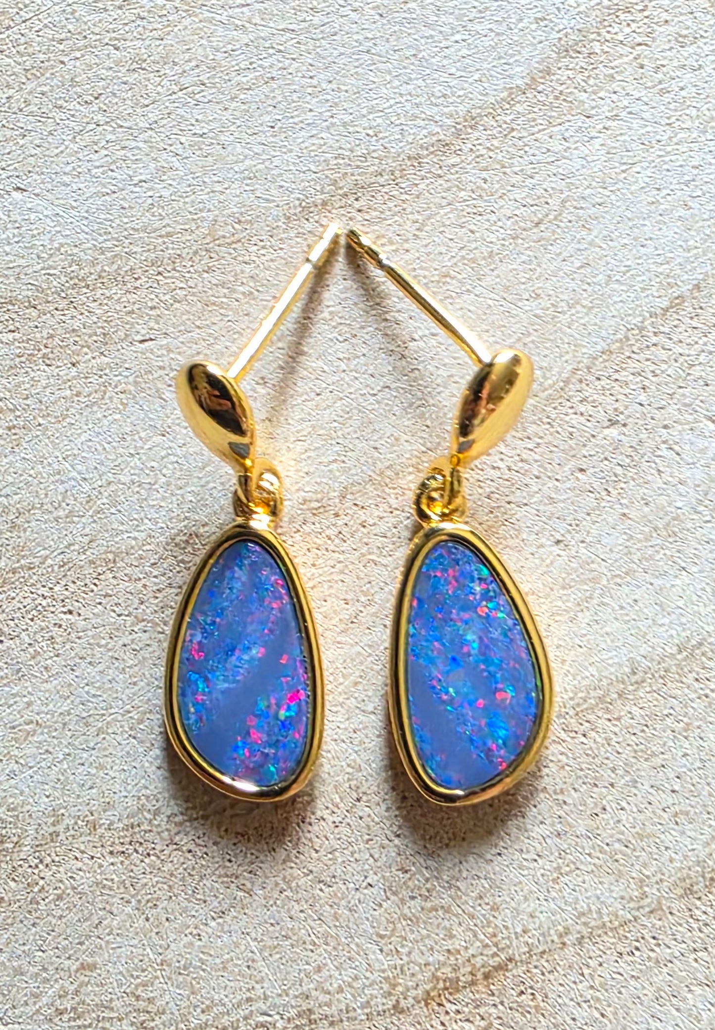 A pair of gold drop earrings with teardrop-shaped blue opal gemstones, displayed on a textured beige surface.